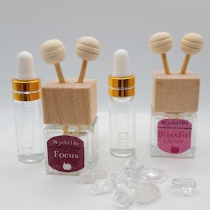 以下が含まれることがあります： 金色の蓋と透明な液体が入ったガラス瓶が3本。それぞれの瓶には、木製のディフューザーと2本の木製のスティックが付いており、ラベルには「WychOils」というテキストと、異なる香りの名前「Focus」、「Blissful Cheer」、「Focus」が書かれています。瓶は、透明な水晶が置かれた白い表面の上に置かれています。