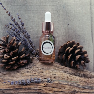 Puede incluir: Una pequeña botella de vidrio con una tapa y un gotero de color oro rosa. La botella está llena de un líquido transparente y contiene botánicos secos. La etiqueta de la botella dice "WyehOils". La botella está sentada sobre un trozo de madera a la deriva con piñas y lavanda.