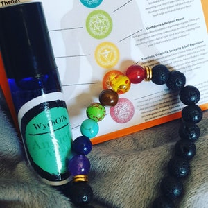 May include: A black lava rock bracelet with seven colourful gemstone beads, including turquoise, green, orange, red, purple, and blue. The bracelet is lying on a white surface with a green and blue bottle labelled "WychOils Anxiety Away" and a chart with seven chakra symbols.