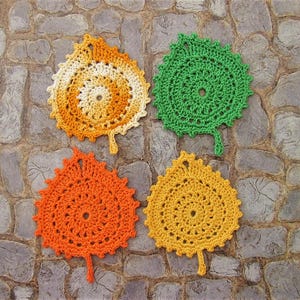 May include: Four crocheted leaf-shaped coasters in orange, yellow, white, and green. The coasters are laid out on a stone surface.