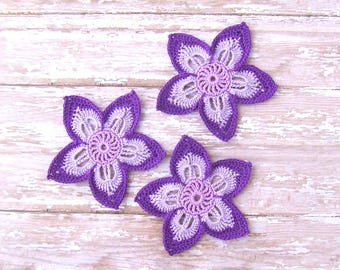May include: Three handmade crochet flowers in shades of purple and white are arranged on a weathered, light-coloured wooden surface. Each flower has five petals and a detailed centre, showcasing the intricate crochet work. The flowers are a charming decorative item.