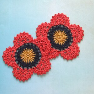 May include: Two handmade, crocheted flower-shaped coasters. The coasters feature a red outer layer, a black middle layer, and a yellow center. The coasters are placed on a light blue surface.
