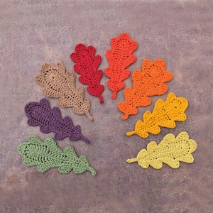 May include: A selection of handmade crochet oak leaves in autumnal hues: red, orange, yellow, green, purple, and brown. The leaves are displayed on a textured purple surface, highlighting the detailed crochet work.
