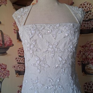 Puede incluir: Vestido de novia color marfil con adornos florales. El vestido presenta un escote cuadrado y mangas cortas. El corpi&ntilde;o est&aacute; adornado con grandes apliques de flores con lentejuelas. El vestido se exhibe en un maniqu&iacute;.