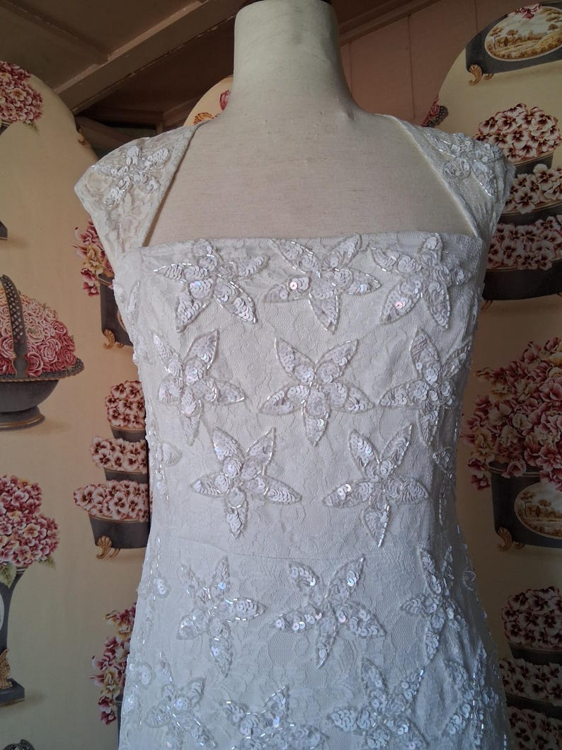 Puede incluir: Vestido de novia color marfil con adornos florales. El vestido presenta un escote cuadrado y mangas cortas. El corpi&ntilde;o est&aacute; adornado con grandes apliques de flores con lentejuelas. El vestido se exhibe en un maniqu&iacute;.
