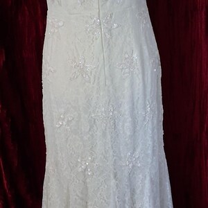 Puede incluir: Vestido de novia de encaje marfil con adornos florales y lentejuelas. La parte trasera del vestido presenta un dise&ntilde;o de ojo de cerradura y cierre de cremallera. El vestido se exhibe en un maniqu&iacute;, mostrando su elegante silueta y detalles intrincados.