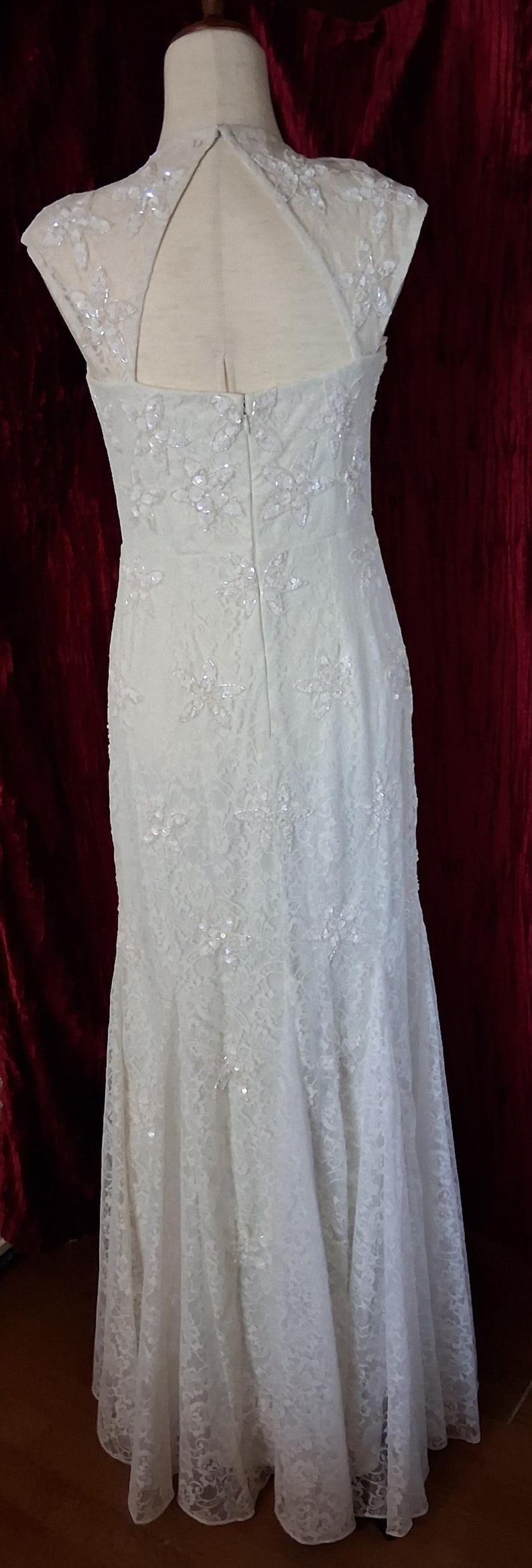 Puede incluir: Vestido de novia de encaje marfil con adornos florales y lentejuelas. La parte trasera del vestido presenta un dise&ntilde;o de ojo de cerradura y cierre de cremallera. El vestido se exhibe en un maniqu&iacute;, mostrando su elegante silueta y detalles intrincados.