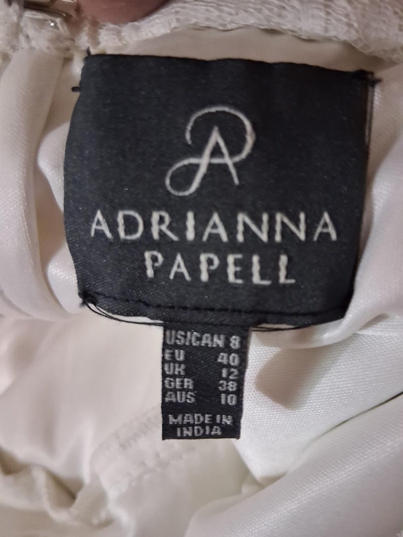 Puede incluir: Primer plano de una etiqueta negra con el texto blanco "ADRIANNA PAPELL". La etiqueta incluye conversiones de tallas para US/CAN, EU, UK, GER y AUS. La etiqueta tambi&eacute;n dice "MADE IN INDIA".