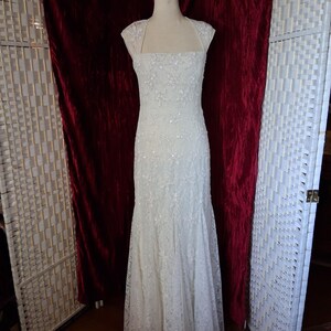 Puede incluir: Un vestido de novia blanco, largo hasta el suelo, con un corpi&ntilde;o ajustado y una falda acampanada. El vestido est&aacute; adornado con encaje floral y lentejuelas. Tiene un escote cuadrado y mangas cortas. El vestido se exhibe en un maniqu&iacute;.