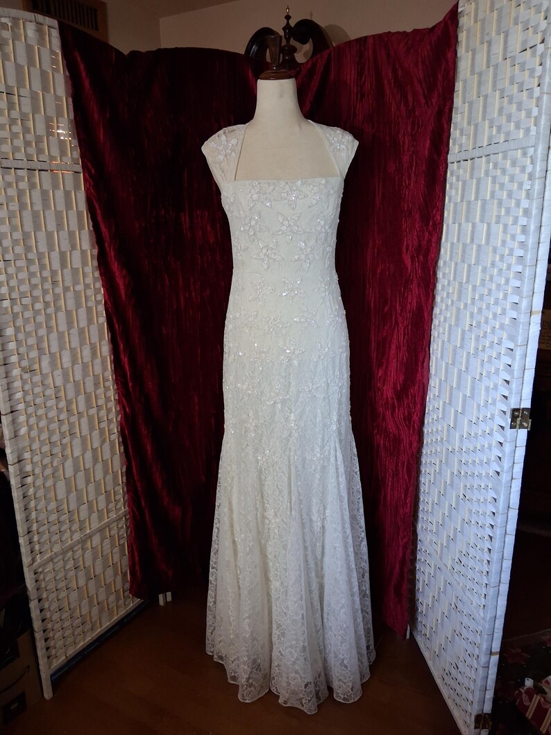 Puede incluir: Un vestido de novia blanco, largo hasta el suelo, con un corpi&ntilde;o ajustado y una falda acampanada. El vestido est&aacute; adornado con encaje floral y lentejuelas. Tiene un escote cuadrado y mangas cortas. El vestido se exhibe en un maniqu&iacute;.