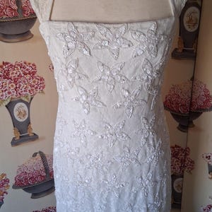 Puede incluir: Vestido de novia color marfil con escote cuadrado y adornos en forma de mariposa. El vestido sin mangas presenta un corpi&ntilde;o ajustado y una falda acampanada. La tela parece ser encaje o un material delicado similar.
