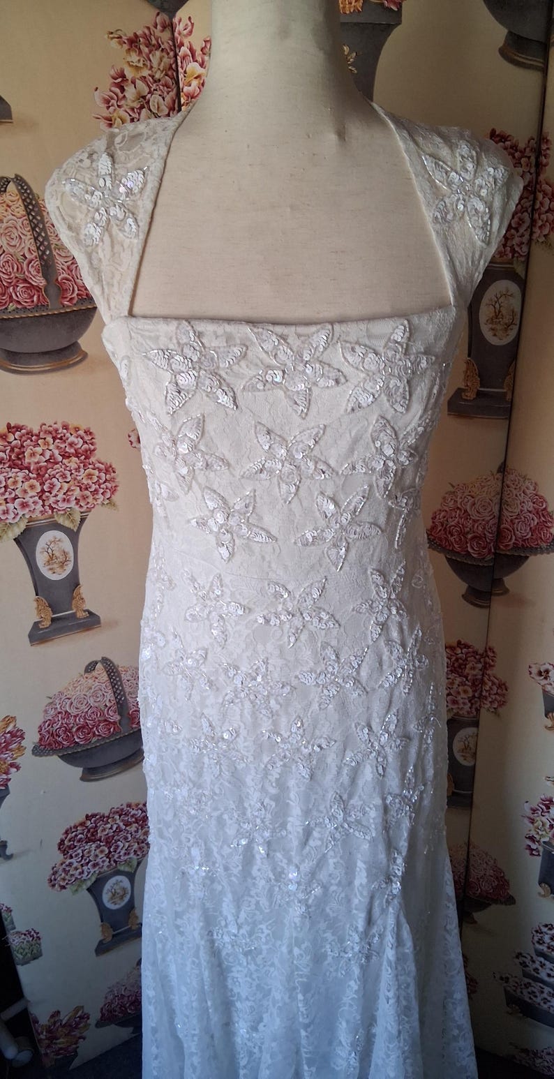 Puede incluir: Vestido de novia color marfil con escote cuadrado y adornos en forma de mariposa. El vestido sin mangas presenta un corpi&ntilde;o ajustado y una falda acampanada. La tela parece ser encaje o un material delicado similar.