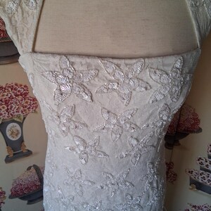 Puede incluir: Cuerpo de vestido de novia color marfil con adornos florales de lentejuelas. El vestido presenta un escote cuadrado y mangas cortas. La tela parece ser encaje o un material delicado similar, con dise&ntilde;o floral.