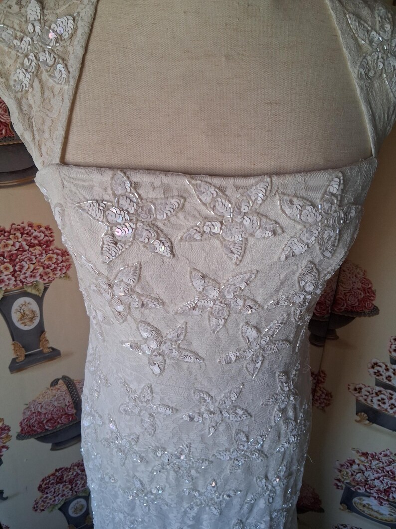 Puede incluir: Cuerpo de vestido de novia color marfil con adornos florales de lentejuelas. El vestido presenta un escote cuadrado y mangas cortas. La tela parece ser encaje o un material delicado similar, con dise&ntilde;o floral.