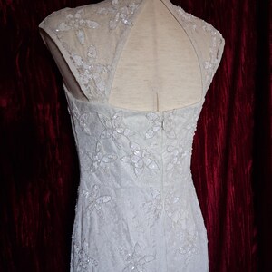Puede incluir: Un vestido claro sin mangas con bordados florales y lentejuelas. El vestido presenta un cuello alto y una espalda con abertura. El vestido se exhibe en un maniqu&iacute; sobre un fondo rojo oscuro.