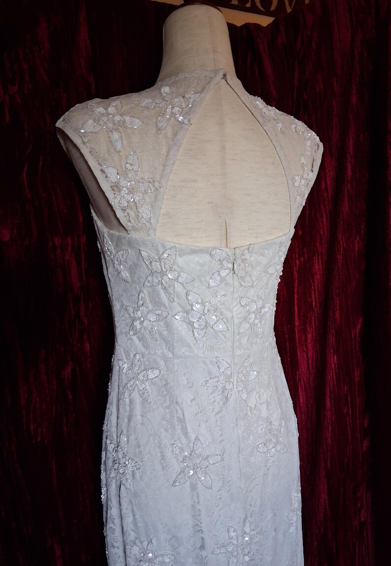 Puede incluir: Un vestido claro sin mangas con bordados florales y lentejuelas. El vestido presenta un cuello alto y una espalda con abertura. El vestido se exhibe en un maniqu&iacute; sobre un fondo rojo oscuro.