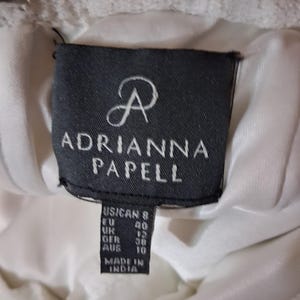 Puede incluir: Primer plano de una etiqueta de vestido blanco con el nombre de la marca "ADRIANNA PAPELL" en texto blanco sobre un fondo negro. La etiqueta incluye conversiones de tallas para tallas de EE. UU., UE, Reino Unido, GER y AUS. El vestido est&aacute; hecho en India.