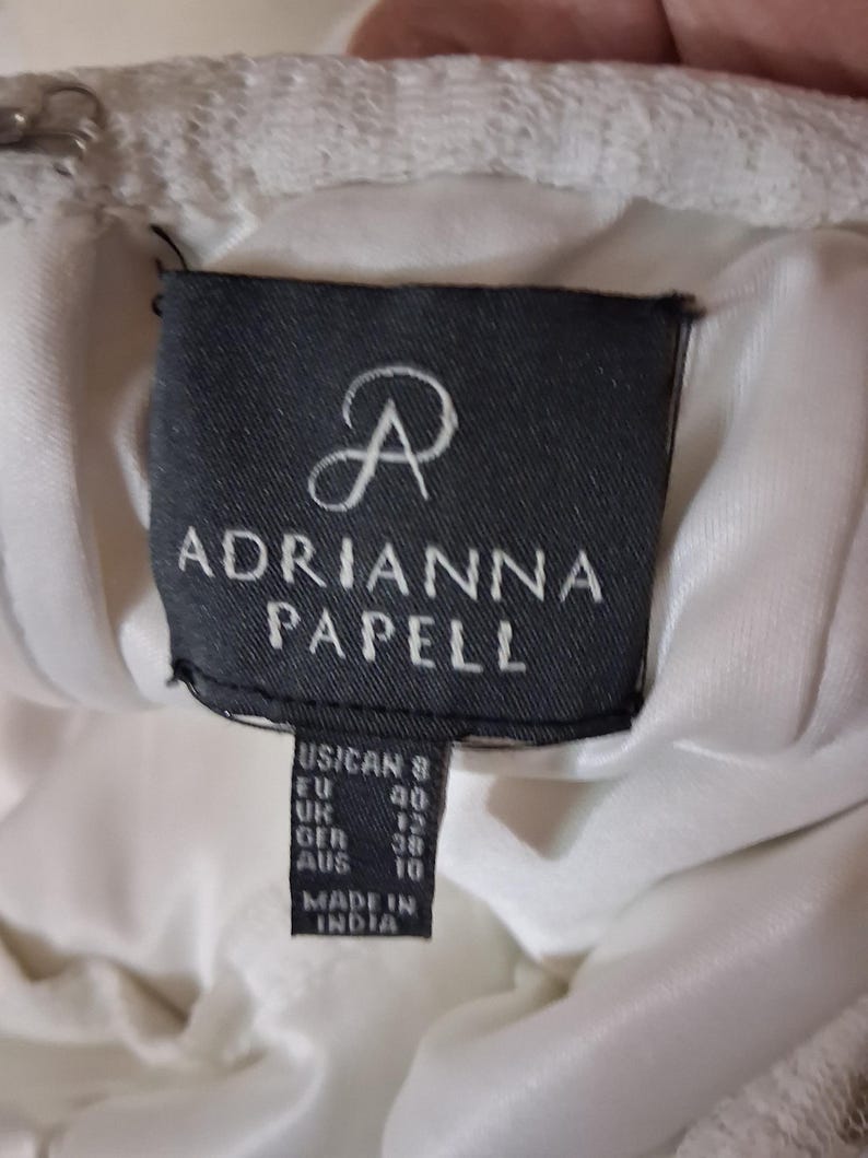 Puede incluir: Primer plano de una etiqueta de vestido blanco con el nombre de la marca "ADRIANNA PAPELL" en texto blanco sobre un fondo negro. La etiqueta incluye conversiones de tallas para tallas de EE. UU., UE, Reino Unido, GER y AUS. El vestido est&aacute; hecho en India.