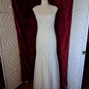 Puede incluir: Un vestido de novia blanco, largo hasta el suelo, con un corpi&ntilde;o ajustado y una falda acampanada. El vestido est&aacute; adornado con encaje floral y lentejuelas. Tiene un escote cuadrado y mangas cortas. El vestido se exhibe en un maniqu&iacute;.