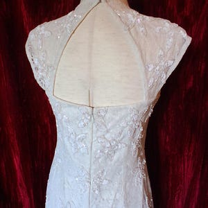Puede incluir: Vestido de novia color marfil con bordados florales y lentejuelas. El vestido presenta un cuello alto y una espalda con abertura. El vestido se exhibe en un maniqu&iacute;. Las palabras "FAITH HOPE" son visibles en el fondo.