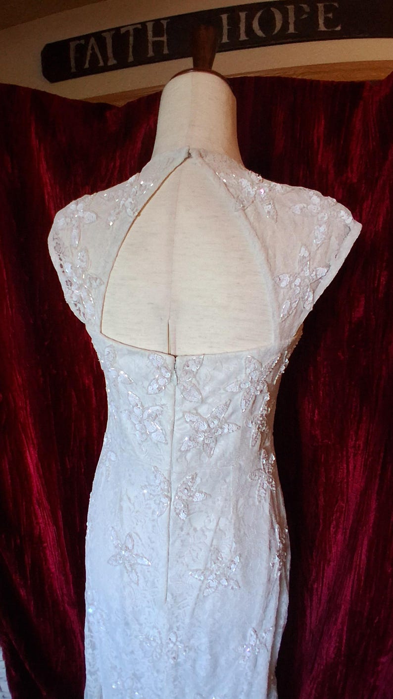 Puede incluir: Vestido de novia color marfil con bordados florales y lentejuelas. El vestido presenta un cuello alto y una espalda con abertura. El vestido se exhibe en un maniqu&iacute;. Las palabras "FAITH HOPE" son visibles en el fondo.