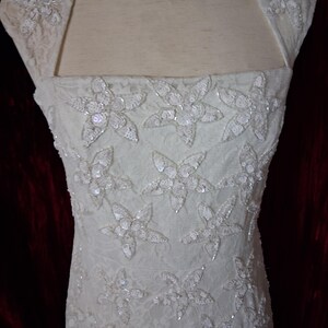 Puede incluir: Vestido de novia color marfil con adornos florales. El vestido presenta un escote cuadrado y mangas cortas. El corpi&ntilde;o est&aacute; adornado con grandes apliques de flores con lentejuelas. La tela parece ser encaje, agregando textura al vestido de novia.