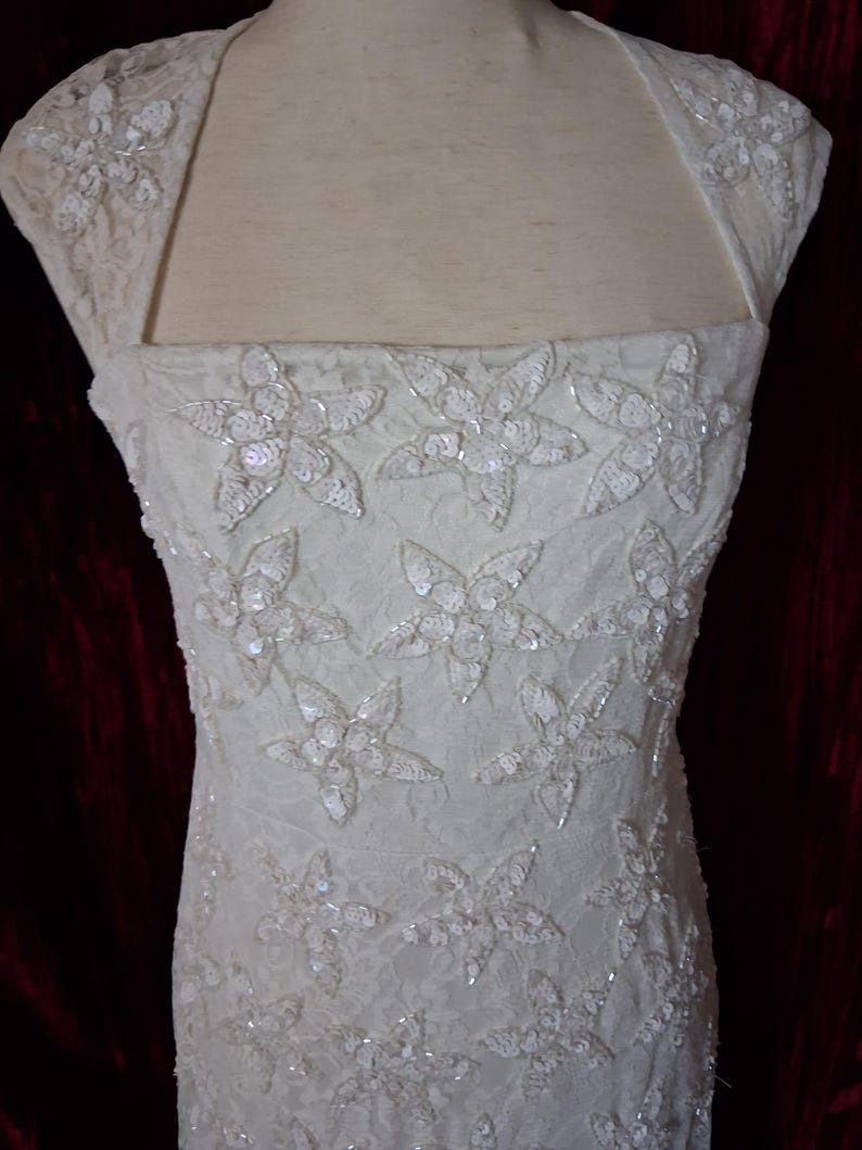 Puede incluir: Vestido de novia color marfil con adornos florales. El vestido presenta un escote cuadrado y mangas cortas. El corpi&ntilde;o est&aacute; adornado con grandes apliques de flores con lentejuelas. La tela parece ser encaje, agregando textura al vestido de novia.