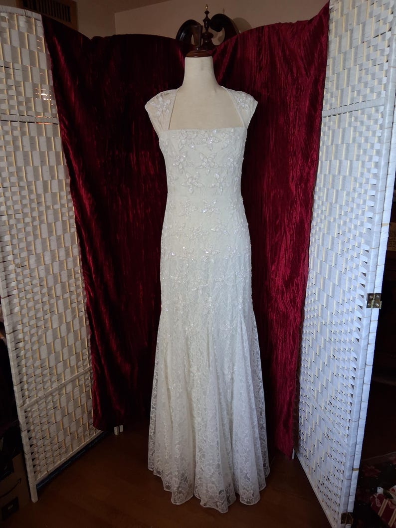 Puede incluir: Un vestido de novia blanco, largo hasta el suelo, con un corpi&ntilde;o ajustado y una falda acampanada. El vestido est&aacute; adornado con encaje floral y lentejuelas. Tiene un escote cuadrado y mangas cortas. El vestido se exhibe en un maniqu&iacute; sobre un fondo de terciopelo rojo.