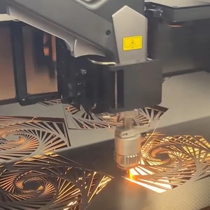 Peut inclure: Une machine de découpe laser découpe avec précision des motifs en spirale complexes dans des feuilles de métal. La buse de la machine émet une lumière orange vif pendant qu'elle travaille, créant des motifs géométriques détaillés.