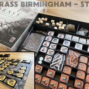 May include: The board game "Brass Birmingham" is displayed with its box and components. The box is grey and black, with the game title prominently featured. Inside a black tray are wooden cylinders, cards, and various game tokens.