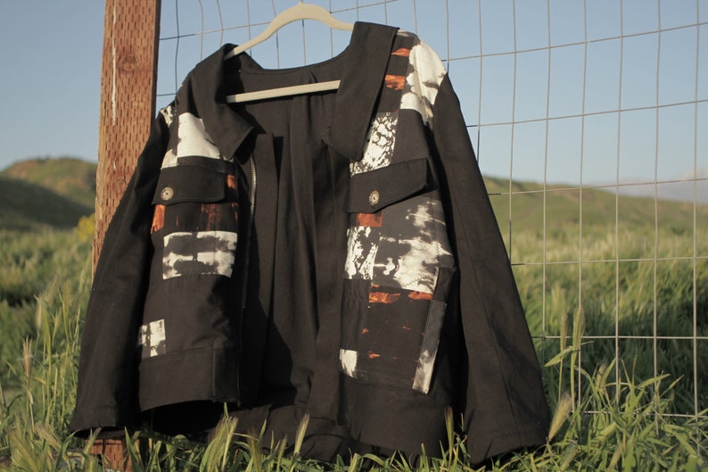 Puede incluir: Una chaqueta vaquera negra con un estampado abstracto blanco y naranja en la parte delantera y trasera. La chaqueta tiene una cremallera y dos bolsillos.