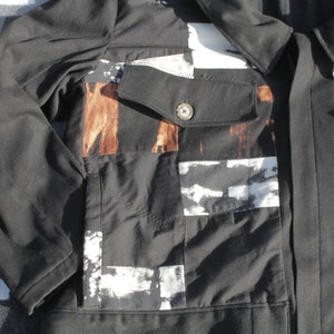 Puede incluir: Una chaqueta negra con un dise&ntilde;o de patchwork con patrones abstractos blancos y marrones. La chaqueta tiene un cierre de bot&oacute;n y un solo bolsillo en el lado izquierdo.