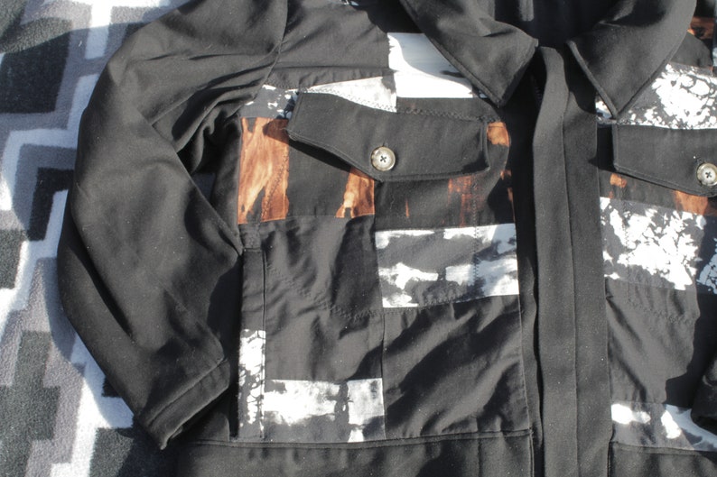 Puede incluir: Una chaqueta negra con un dise&ntilde;o de patchwork con patrones abstractos blancos y marrones. La chaqueta tiene un cierre de bot&oacute;n y un solo bolsillo en el lado izquierdo.