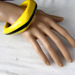 Op de afbeelding: Een dikke, grove armband met een felgele basiskleur en een opvallende, effen zwarte streep die horizontaal rond het midden van het sieraad loopt. De armband heeft een gladde, glanzende afwerking en een afgerond, gebogen buitenprofiel.