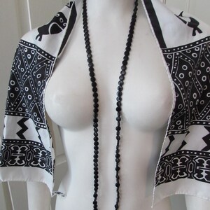 May include: A black beaded necklace hangs over a white mannequin. A white scarf with black geometric and animal print is draped over the mannequin's shoulders. The scarf has a black border.