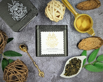 Puede incluir: Una lata negra con un escudo dorado y el texto "Milk Oolong" en la parte delantera. La lata está rodeada de varios objetos, incluyendo un colador de té dorado, una bola de madera y una pequeña cuchara dorada.