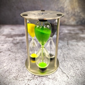 Op de afbeelding: Een zilveren zandloper met drie kamers gevuld met groen, geel en zwart zand. De zandloper is gelabeld "3 minute light".