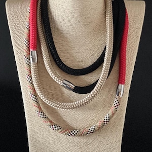 May include: A set of four layered necklaces made of rope. The necklaces are in black, beige, red, and a beige and black plaid pattern. The necklaces are on a mannequin.