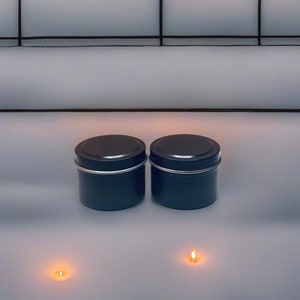 May include: Two black metal candle tins with lids, sitting on a white surface. Two small lit candles are in front of the tins.
