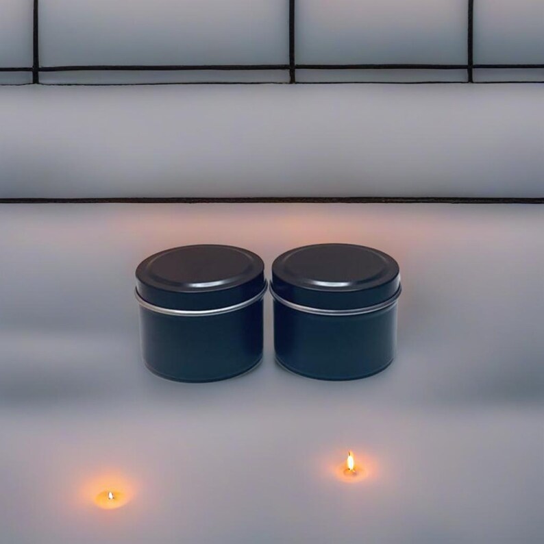 May include: Two black metal candle tins with lids, sitting on a white surface. Two small lit candles are in front of the tins.
