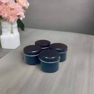 May include: Four black metal candle tins with lids. The tins are arranged in a square shape on a light wood surface.