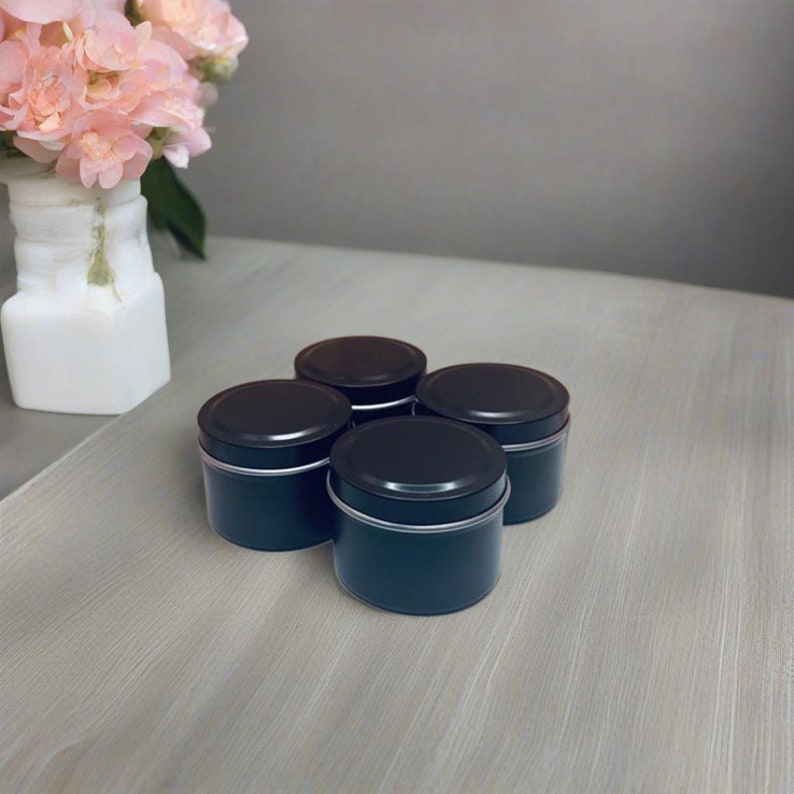 May include: Four black metal candle tins with lids. The tins are arranged in a square shape on a light wood surface.