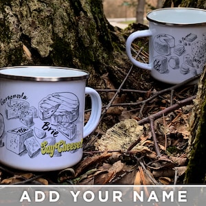 May include: Two white enamel mugs with black and yellow illustrations of different types of cheese. The mugs have black rims and handles. The text on the mugs reads "Gorgonzola", "Brie", "Say Cheese!" and "Cheddar", "Parmesan", "Swiss".