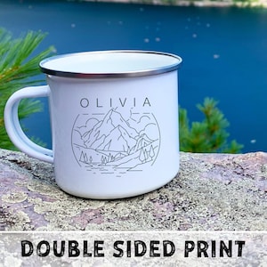 May include: White enamel camping mug with a black outline of a mountain scene and the name "Olivia" printed on the front. The mug is sitting on a rock with a lake in the background.