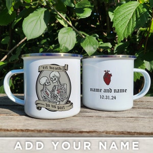 May include: Two white enamel mugs with a black and white skeleton couple design on one mug and a red heart with the text "name and name 10.31.24" on the other mug.