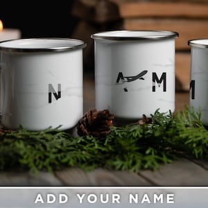 May include: Three white enamel mugs with silver rims, each displaying a different letter in black. The middle mug features a stylized airplane graphic. The mugs are arranged on a wooden surface with greenery and a pinecone, with a blurred background.