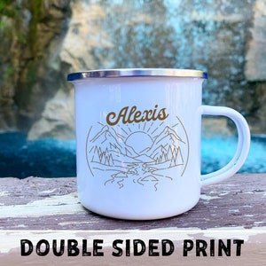 May include: White enamel camping mug with a silver rim and handle. The mug features a mountain and sun design with the name "Alexis" in gold lettering. The words "DOUBLE SIDED PRINT" are printed at the bottom.