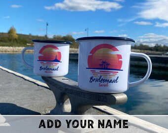 May include: Two white enamel mugs with black rims and handles. Each mug features a sunset design with the word "Bridesmaid" and a name. One mug has a lighthouse, the other has palm trees. The text "ADD YOUR NAME" is at the bottom.