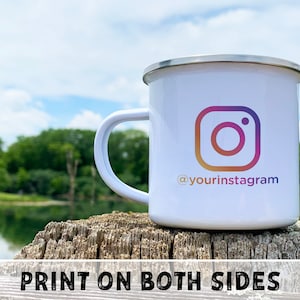 May include: White enamel camping mug with a colorful Instagram logo and the text "@yourinstagram" printed on the side.  The mug is sitting on a wooden surface.  Print on both sides.