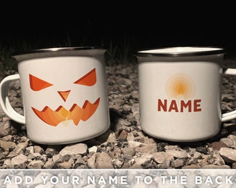 May include: Two white enamel mugs with silver rims. One mug features an orange jack-o'-lantern face with a candle, the other has a candle design and the word "NAME" in orange. The mugs have white handles and are on a rocky surface.
